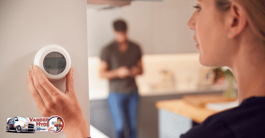 A woman holding a manual thermostat on the wall with a man leaning on a counter in the background